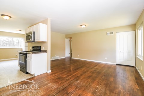A spacious living room with wood flooring and a kitchen area in the background.