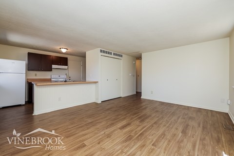 A room with a wooden floor and a kitchen area with a refrigerator, microwave, and cabinets.