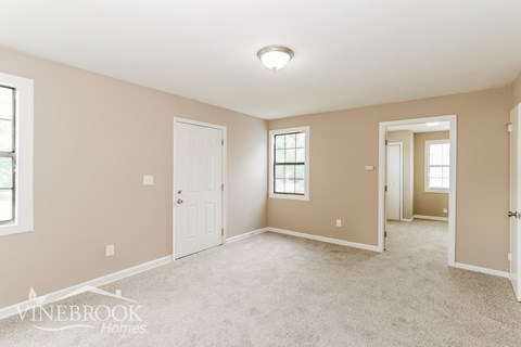 A spacious room with beige walls and carpet flooring, with a window on the left wall and a door on the right wall.