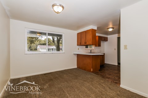 A room with a carpeted floor, a window, and a kitchenette area with cabinets and a countertop.