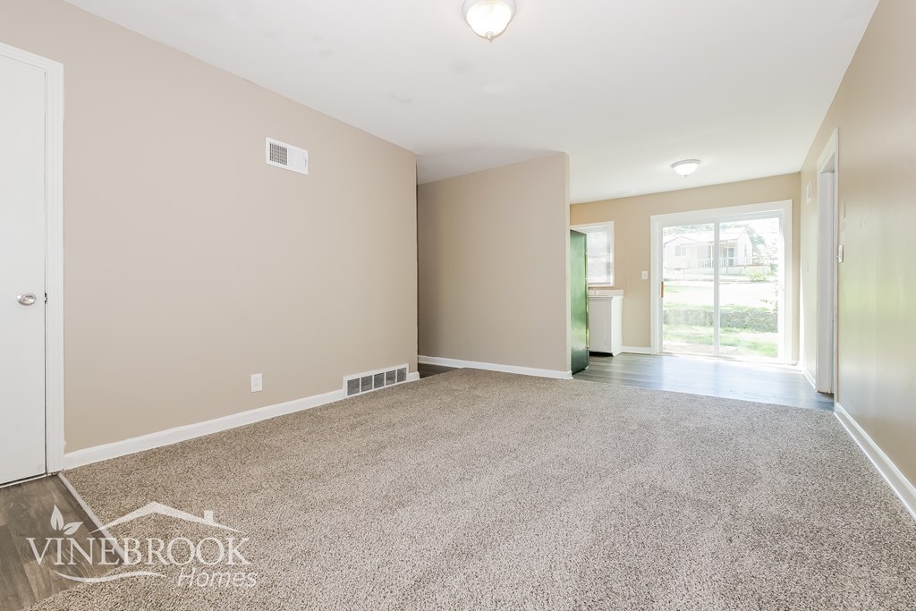 A room with beige walls and carpet flooring, with a door on the left and a window on the right.