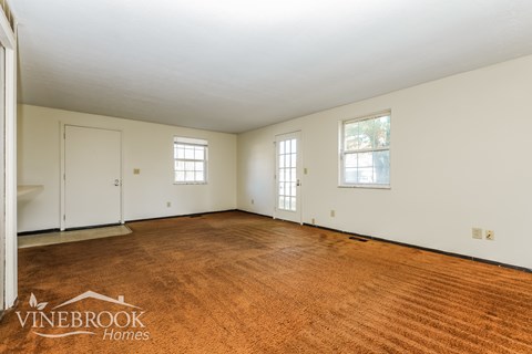 A room with a wooden floor and white walls, with a door on the left and two windows on the right.