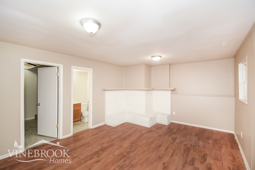 A room with wood flooring and white walls, with a door open to a bathroom.