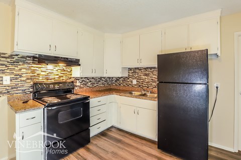 A black fridge stands in a kitchen with white cabinets and a stone backsplash.