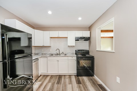 A kitchen with a black fridge and black dishwasher with white cabinets and a wooden floor.