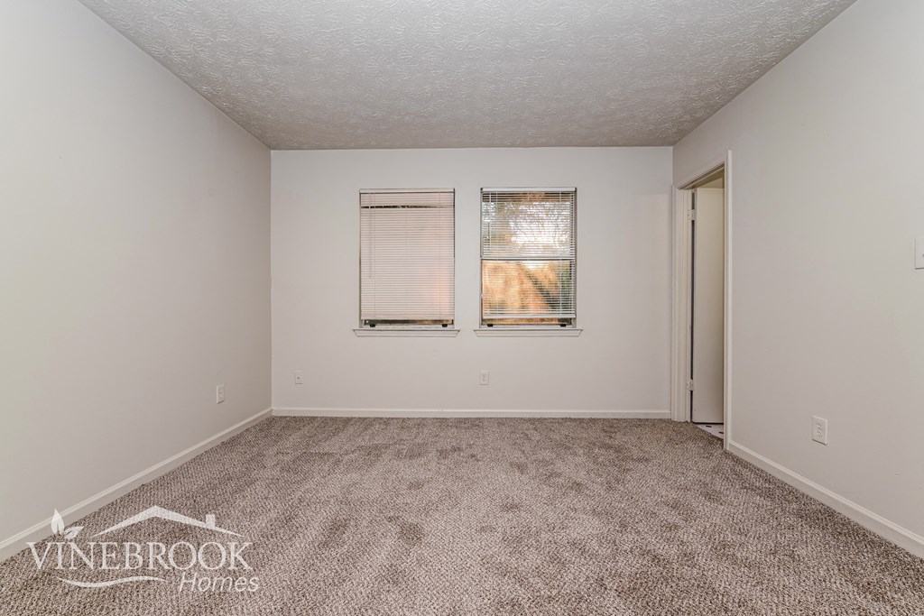 a spacious living room with carpet and two windows