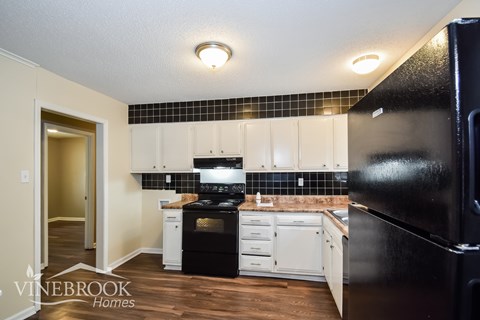 A black refrigerator is in the foreground of a kitchen with white cabinets and black and white tiles.