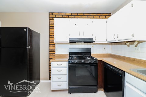 A black refrigerator is in a kitchen with white cabinets and a brick wall.