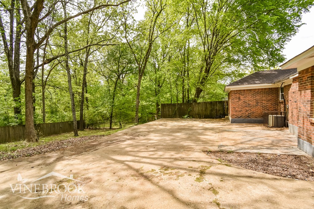 A dirt driveway leads to a small brick building with a fence and trees in the background.