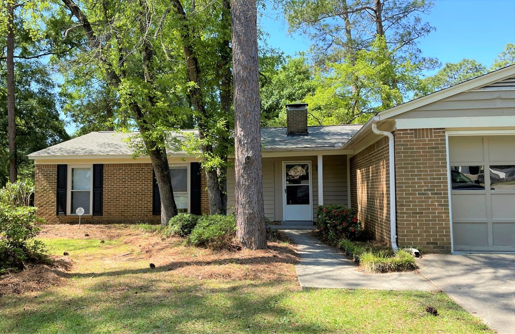 A house with a brown brick exterior and a white door is surrounded by greenery.
