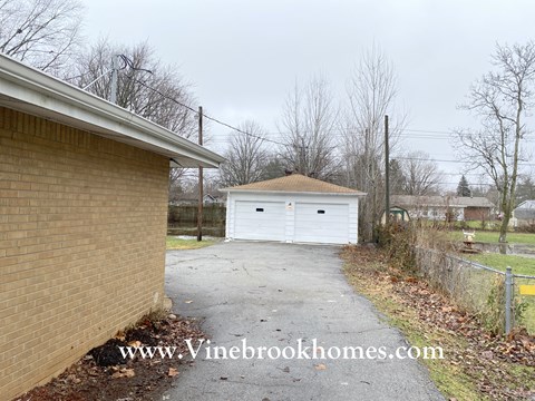 A white garage is situated in a driveway between a brick building and a fence.