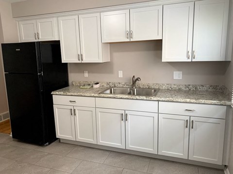 A black refrigerator stands next to a kitchen with white cabinets.