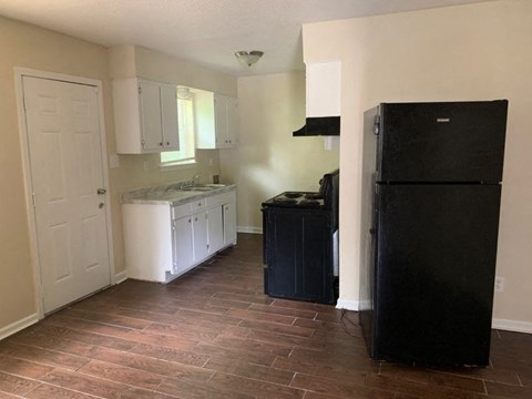 A black refrigerator stands in a kitchen with white cabinets and a wooden floor.