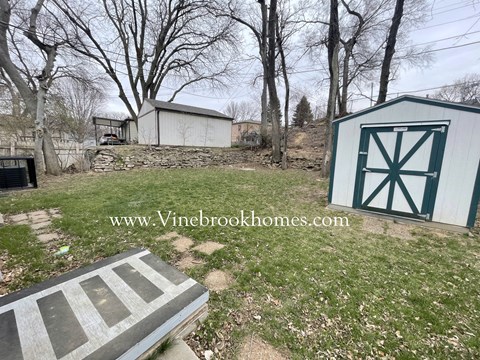 A white shed with a green door is situated in a grassy area with a stone wall and a tree in the background.