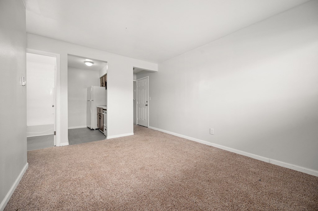 a living room with white walls and carpet and a kitchen