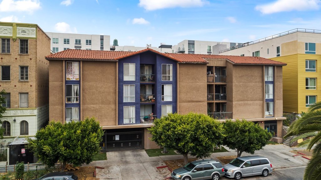 A building with a red tile roof is in the middle of a street with cars parked in front.