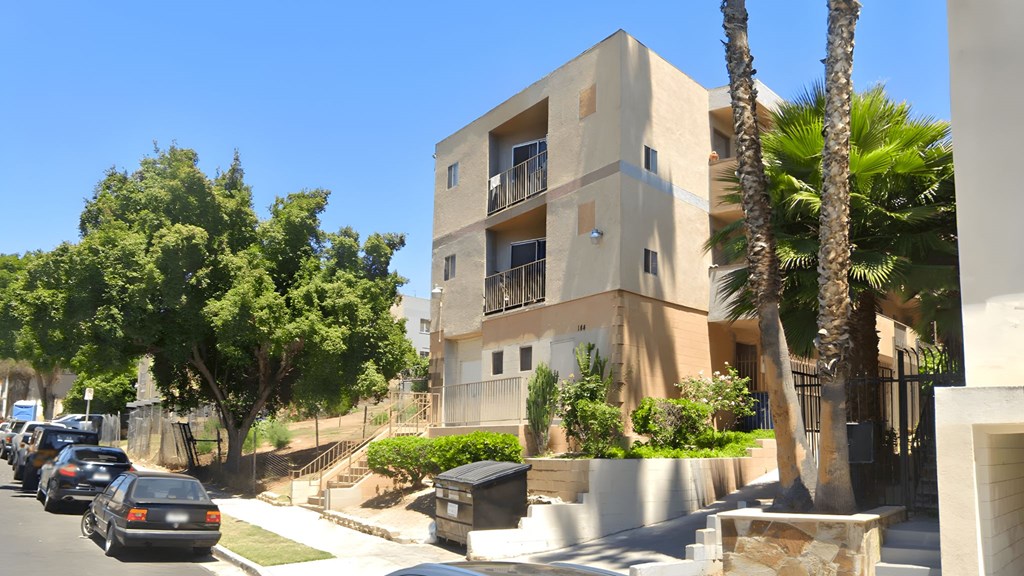 A modern building with a balcony is surrounded by trees and parked cars.
