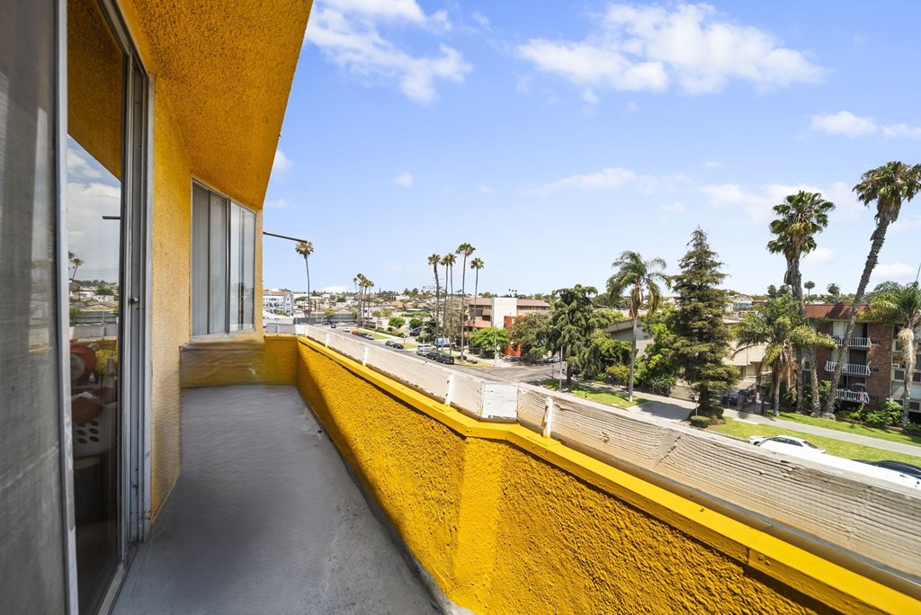 A yellow building with a balcony overlooking a street with palm trees.