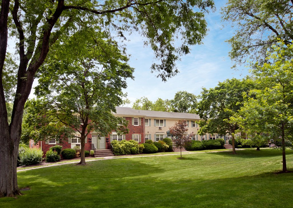 A large red brick building with a white door and windows is surrounded by green trees and grass. at Rivercrest Apartments, New Jersey, 08854