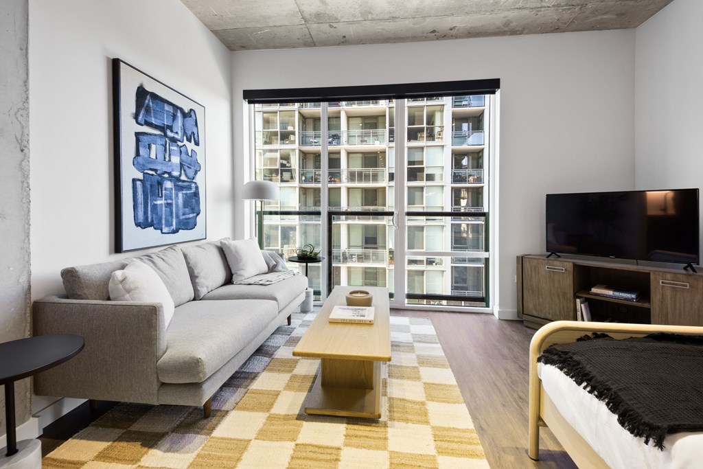 A living room with a grey couch, a yellow coffee table, a black and white checkered rug, a black bed, a black TV, and a large window with a view of a building.