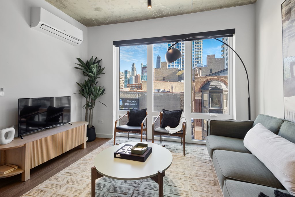 A living room with a television, a couch, a chair, a table, and a window with a view of the city.