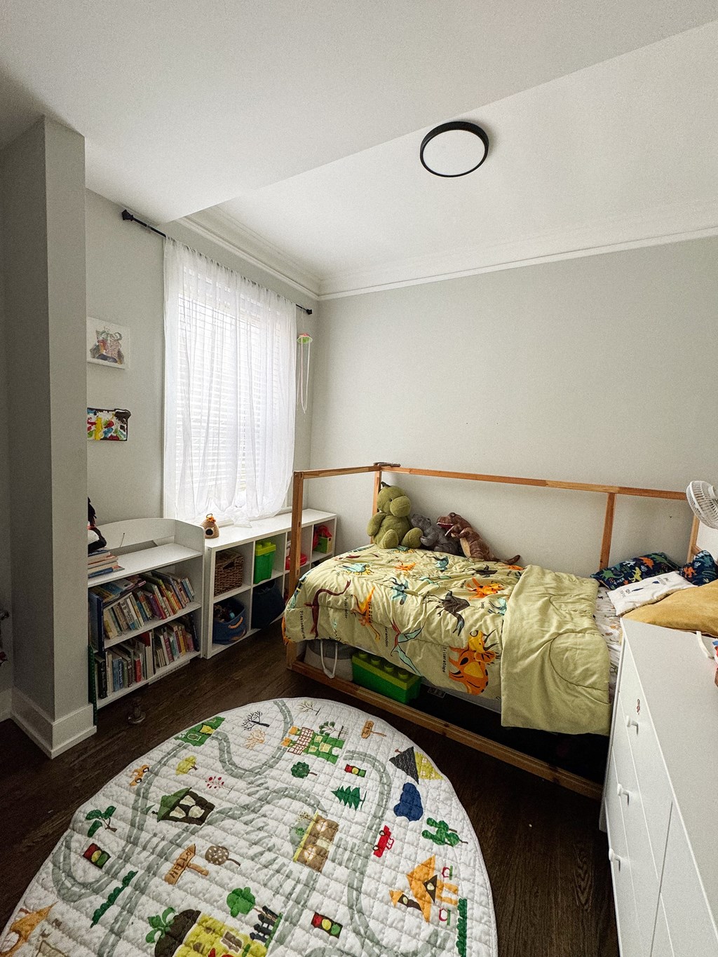A bedroom with a bed, a round rug, a bookcase, and a window with curtains.