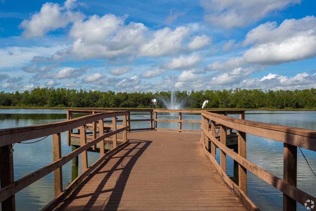 A wooden pier extends into a body of water with a fountain in the distance.