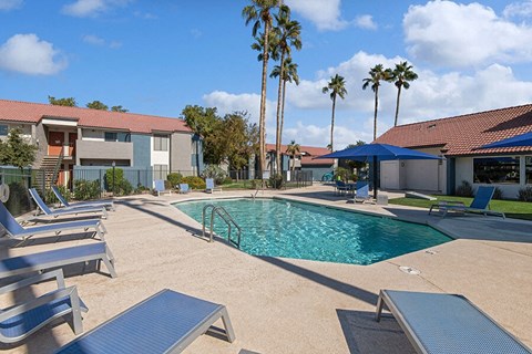 Avondale, AZ, Apartments Near Goodyear Ballpark - The Peaks on 4th - A Swimming Pool With Lounge Chairs, Blue Umbrellas, Palm Trees, And Apartment Buildings.