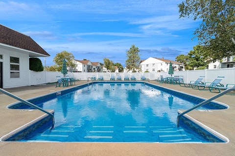 Apartments for rent in Marshfield, MA - Village At Marshfield - Resort-Style Outdoor Pool With Blue Water, Entry Handrails, Lounge Chairs, And Umbrellas Inside White Fenced Courtyard.