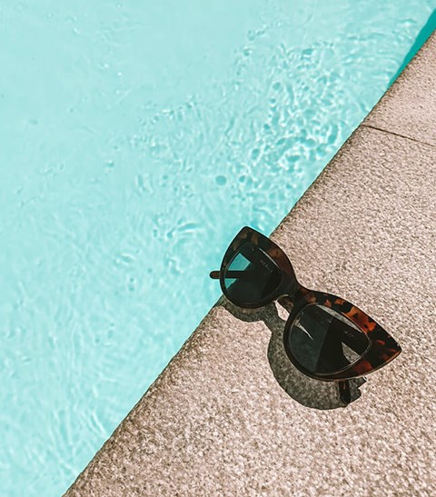 A pair of sunglasses with a black frame and orange accents are resting on the edge of a pool.