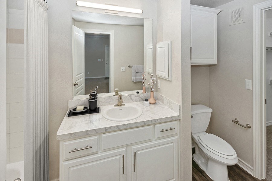 Model Bathroom with White Cabinets and Wood-Style Flooring at Belmont at Duck Creek Apartments in Garland, TX.