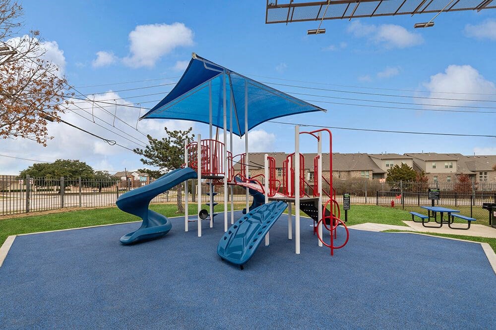 Community Playground with Slide and Blue Canopy at Bridges at Oakbend Apartments in Lewisville, TX.