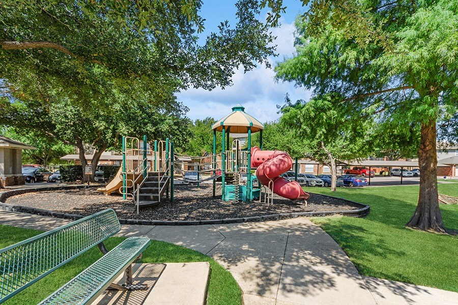 Community Playground with Two Slides and Bench Area at Belmont at Duck Creek Apartments in Garland, TX.