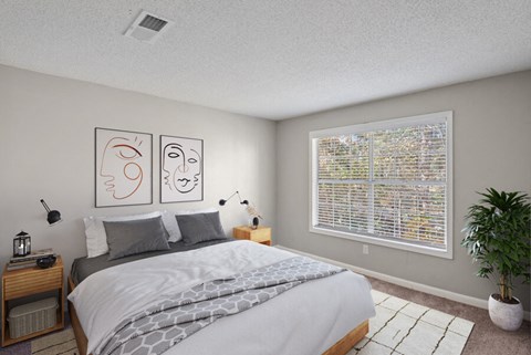 East Charlotte, NC, Apartments Near Albemarle Road Elementary - Woodland Estates - A Bedroom With A Large Bed, Wall Art, Wood Nightstands With Lamps, A Large Window, And A Potted Plant.