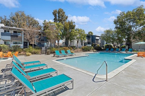 Apartments in El Cajon, CA - Colonnade at Fletcher Hills - Resort-Style Swimming Pool with Turquoise Lounge Chairs, Pool Steps and Handrail, Spacious Deck, and Apartment Buildings Under Blue Sky.