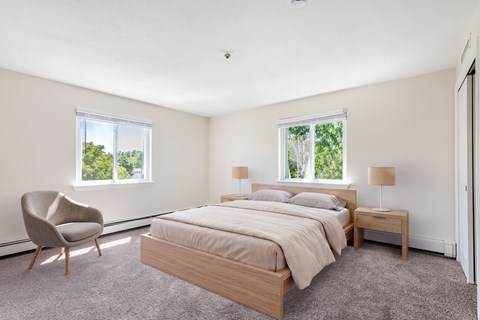 A bedroom with a bed, chair, and two windows.