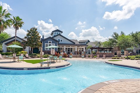 A swimming pool in front of a house with a patio and chairs.