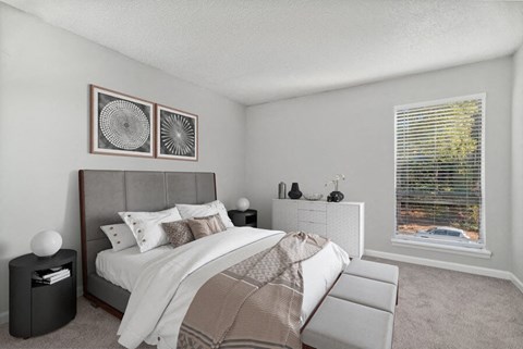 One-Bedroom Apartments in Raleigh, NC - Bridges at North Hills - A Bedroom With Carpet Flooring, A Large Bed, Matching Nightstands, Framed Wall Art, A Dresser, And A Large Window.