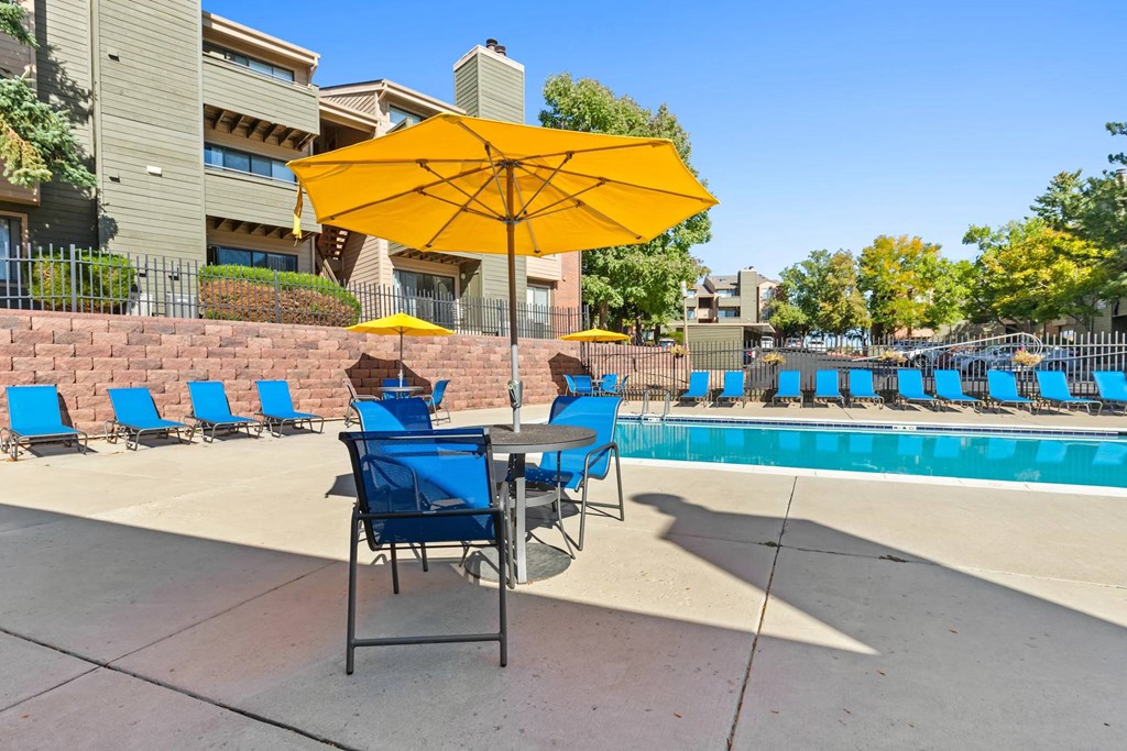 A yellow umbrella shades a table with blue chairs by a pool.