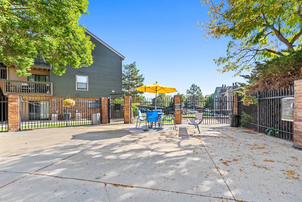 A playground with a yellow umbrella and a blue slide is surrounded by a fence and a building.