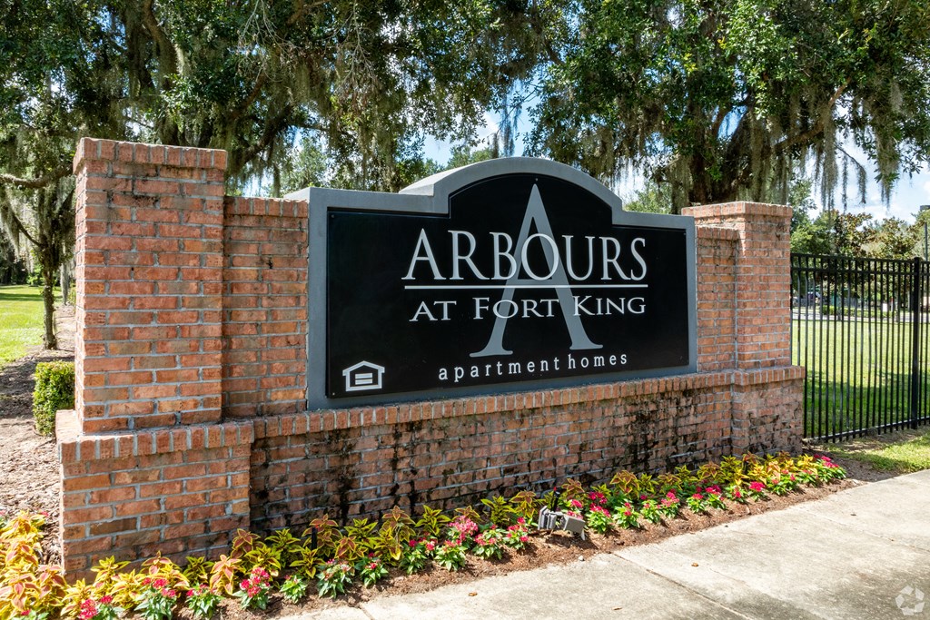 A sign that says Arbours at Fort King stands in front of a brick wall.