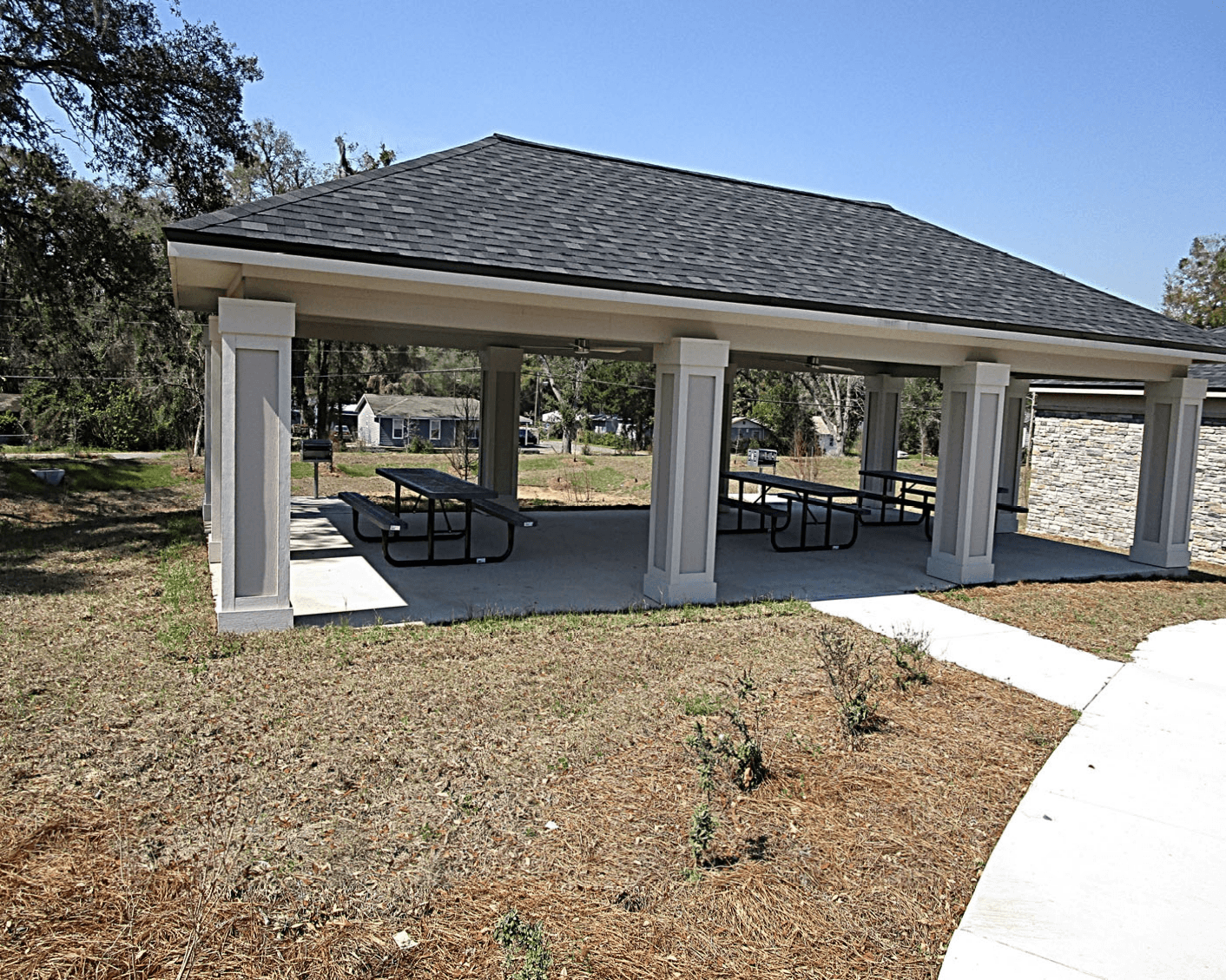 A pavilion with a black roof and white pillars is surrounded by a grassy area with a few small plants.