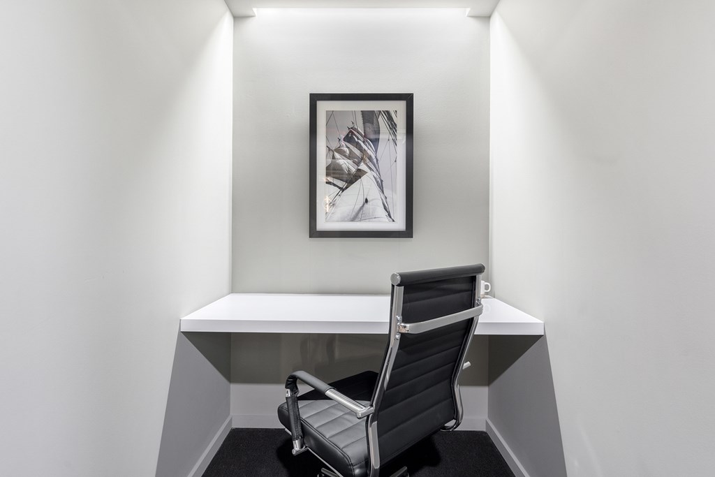 A black and white photo is hanging on a white wall above a white desk with a black chair in front of it.