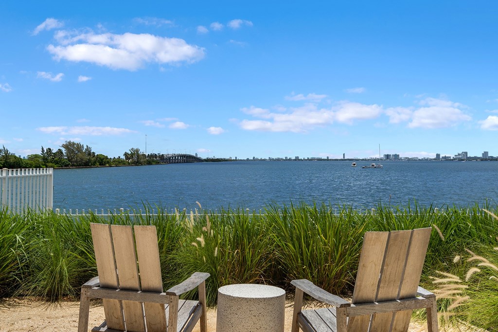 Two chairs and a table are on a beach with a body of water and a city in the background.