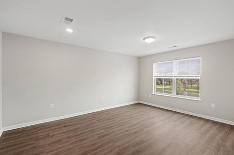 A spacious room with wooden flooring and a window showing a view of trees and a road.