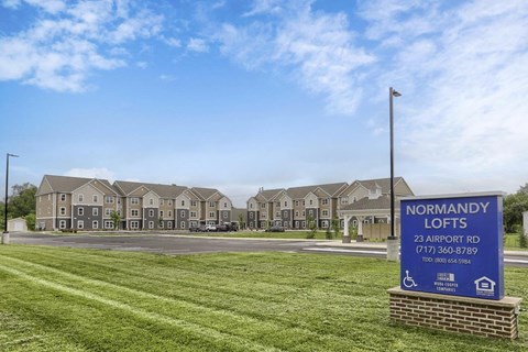 A sign for Normandy Lofts is in front of a building with a grassy area in the foreground.