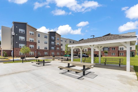 A sunny day at a park with picnic tables and benches in front of apartment buildings.