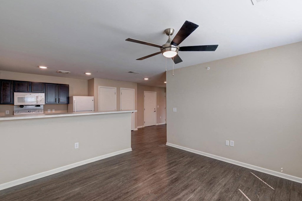 an empty living room with a ceiling fan and a kitchen in the background