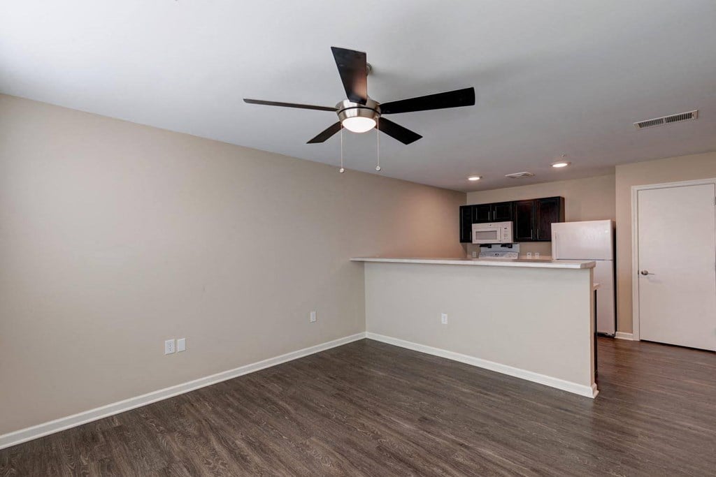 an empty room with a ceiling fan and a kitchen in the background