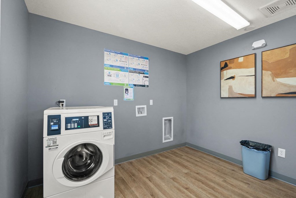 A washing machine is in a room with a blue trash can and two paintings on the wall.